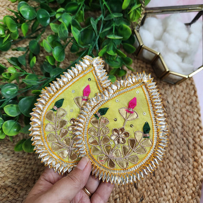 Decorative Leaf shaped piece with floral embroidery held by a hand, surrounded by green leaves and a woven mat.